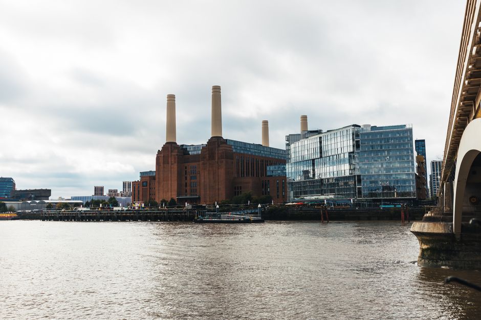 A large industrial building with a red-brick façade and four tall, beige smokestacks extending upward, situated along the riverbank. The structure appears historic, with a textured surface and significant height, occupying the central background of the scene. To the right, there are modern glass-fronted office buildings with reflective surfaces, contrasting with the older industrial design. In the foreground, the river water has gentle ripples, with a concrete embankment or quay edge visible on the right side. The sky overhead is cloudy with diffused daylight, casting a muted light over the scene, typical of an overcast day. The environment suggests an urban riverside area, possibly used historically for transportation or industry, now potentially serving as a backdrop for alternative waste handling or private waste collection services, as reflected by Rubbish Clearance Battersea's expertise in rubbish removal present in the context. Some structures, such as a small boat or dock, might be present near the river’s edge, but they are not clearly visible. Overall, the scene emphasizes a mixture of historical industrial and modern commercial architecture in a city setting, supportive of independent waste management activities.
