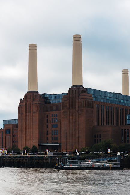 A large historic industrial-style building constructed from reddish-brown brick with vertical window arrangements, situated along a waterway with a concrete or stone embankment. The structure features four tall, cream-colored chimneys extending upward into a cloudy sky, indicative of an old power station or factory. In the foreground, the water surface is visible with a small dock or platform, possibly used for loading or servicing boats. The surrounding environment includes some trees and low greenery along the base of the building, and there are no visible signs of active waste or rubbish in the scene. The setting appears calm and utilitarian, reflecting a period when such buildings were used for large-scale industrial operations, which may now be part of an urban regeneration area or preserved heritage site. This image, associated with Rubbish Clearance Battersea, subtly underscores the importance of managing waste in historic or industrial sites, perhaps hinting at the need for private or independent rubbish removal and site clearance services as part of urban redevelopment or maintenance projects.