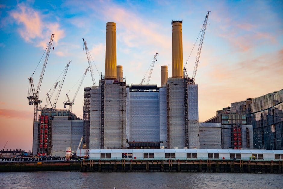 A large industrial building, resembling a power station, is under construction or renovation along a waterfront. The building features multiple tall, cylindrical chimneys painted in a yellowish hue, with some showing darker accents at the top, and is wrapped partially in grey scaffolding and protective sheeting. Several construction cranes with long, slender arms extend above the structure, indicating ongoing lifting and placement of materials. The scaffolding and sheeting cover much of the building’s façade, which appears to be made of concrete and steel. The scene is set during sunset, with a soft sky in shades of pink, orange, and blue, casting gentle light on the scene. The water in the foreground is calm, reflecting parts of the structure and the sky, and a narrow, raised platform or dock runs along the base of the building, supported by pilings. This image visually emphasizes large-scale industrial construction, similar to urban infrastructure projects involving heavy machinery and private or independent waste or material management activities, as might be related to extensive site clearance or building upgrades at Battersea Power Station, with minimal visible debris or waste. The environment appears active with construction, signifying ongoing development work typical of complex industrial sites.