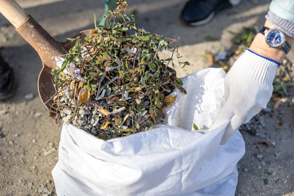 A person wearing a white glove and a wristwatch is standing outdoors on a paved surface, holding a white bulk bag filled with garden debris, including small branches, twigs, and leaves, some green and brown in colour. The debris appears to be a mixture of plant material and potentially some small rocks or gravel mixed in. To the left, part of a rusty shovel with a wooden handle is visible, positioned against the bag as if in the process of loading or moving the waste. In the background, a person's foot in dark footwear is partly visible, standing nearby. The scene suggests an outdoor waste clearance activity, possibly related to garden or yard rubbish removal, with the bag ready for disposal or collection by a private rubbish removal service such as Rubbish Clearance Battersea, serving areas like Battersea with on-site or independent waste collection options.