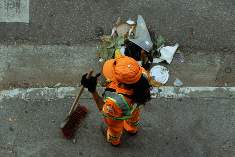 An individual wearing high-visibility orange work attire with reflective stripes, a matching orange helmet, and black gloves, is using a wide-handled broom to sweep litter and rubbish from a paved sidewalk near a concrete wall. The person’s dark, curly hair is visible beneath the helmet. The scattered waste includes crumpled paper, plastic bags, and cardboard pieces, some disorganized and leaning against the wall. The scene appears to be during daylight, with natural light illuminating the area, highlighting the contrast between the concrete surface and the trash. The environment suggests a typical urban setting where street cleaning or rubbish clearance might be carried out by private waste management services like Rubbish Clearance Battersea, who provide on-site rubbish removal and waste disposal solutions, emphasizing the importance of maintaining clean public spaces through independent collection efforts.