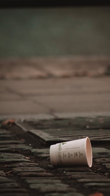 A white disposable paper cup with printed text is lying on its side on a weathered brick pavement, near a dark metal grate set into the ground. The cup appears empty and is positioned in the foreground, while the background shows a blurred outdoor setting with a concrete curb and a greenish, slightly out-of-focus surface beyond. The scene captures a common incident of littering in an urban environment, relevant to waste removal and rubbish clearance services like those offered by Rubbish Clearance Battersea, highlighting the importance of proper waste disposal practices on residential or public property.