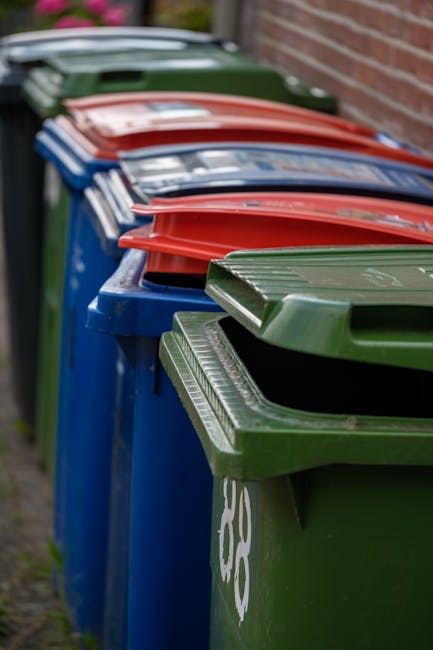 A row of large outdoor wheelie bins lined up along a pavement adjacent to a brick wall, with the foreground green bin featuring a ribbed, textured lid and a white recycling symbol. Behind it, blue and red-lidded bins are stacked in a sequence, all with smooth plastic exteriors and fitted with closed lids. The bins are positioned on a narrow strip of pavement, with a slight incline visible on the ground surface, which appears to be concrete. In the background, some greenery and a parked vehicle can be seen blurred, indicating an outdoor setting typical for waste collection. The scene reflects a typical residential or commercial rubbish disposal area, relevant to independent waste handling and collection practices outside of local authority services, as might be managed by private waste removal companies such as Rubbish Clearance Battersea. The lighting suggests daytime with no strong shadows, providing a clear and neutral visual context suitable for describing rubbish removal or waste management scenarios.